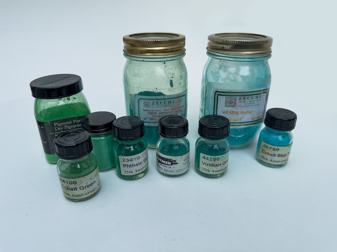 An array of dry green pigments in jars and bottles. Some say viridian, others cobalt green, and a few other pigments. Two jars of phthalo green are shown but neither one is Phthalo Green Blue Shade